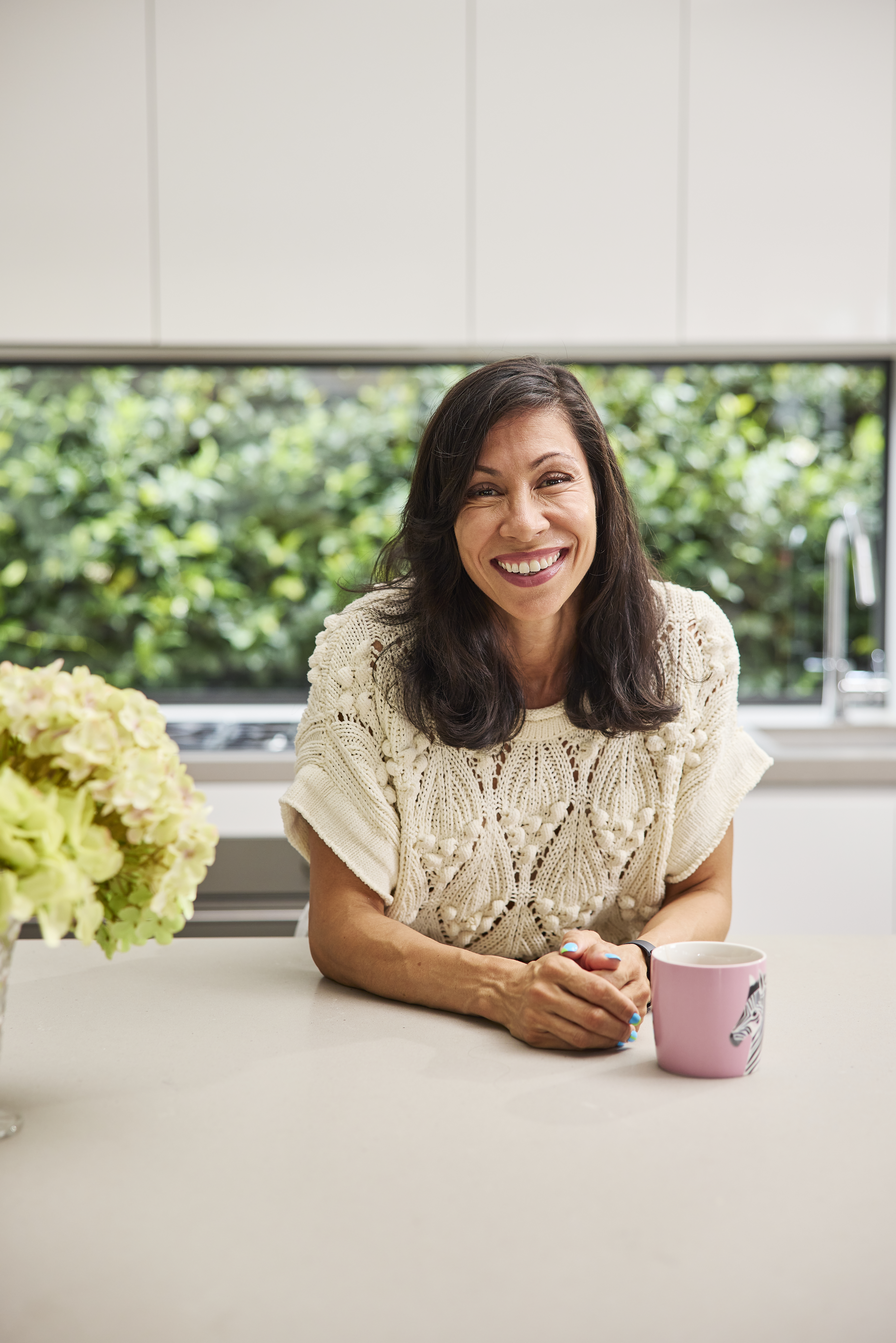 Ilse Martyns is leaning over an island bench in a modern kitchen, smiling. Beside her is a cup of tea that's stewing and a full vase of hydrangeas. Ilse has long, dark hair and is wearing a white knitted top.