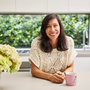 Ilse Martyns is leaning over an island bench in a modern kitchen, smiling. Beside her is a cup of tea that's stewing and a full vase of hydrangeas. Ilse has long, dark hair and is wearing a white knitted top.