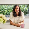 Ilse Martyns is leaning over an island bench in a modern kitchen, smiling. Beside her is a cup of tea that's stewing and a full vase of hydrangeas. Ilse has long, dark hair and is wearing a white knitted top.