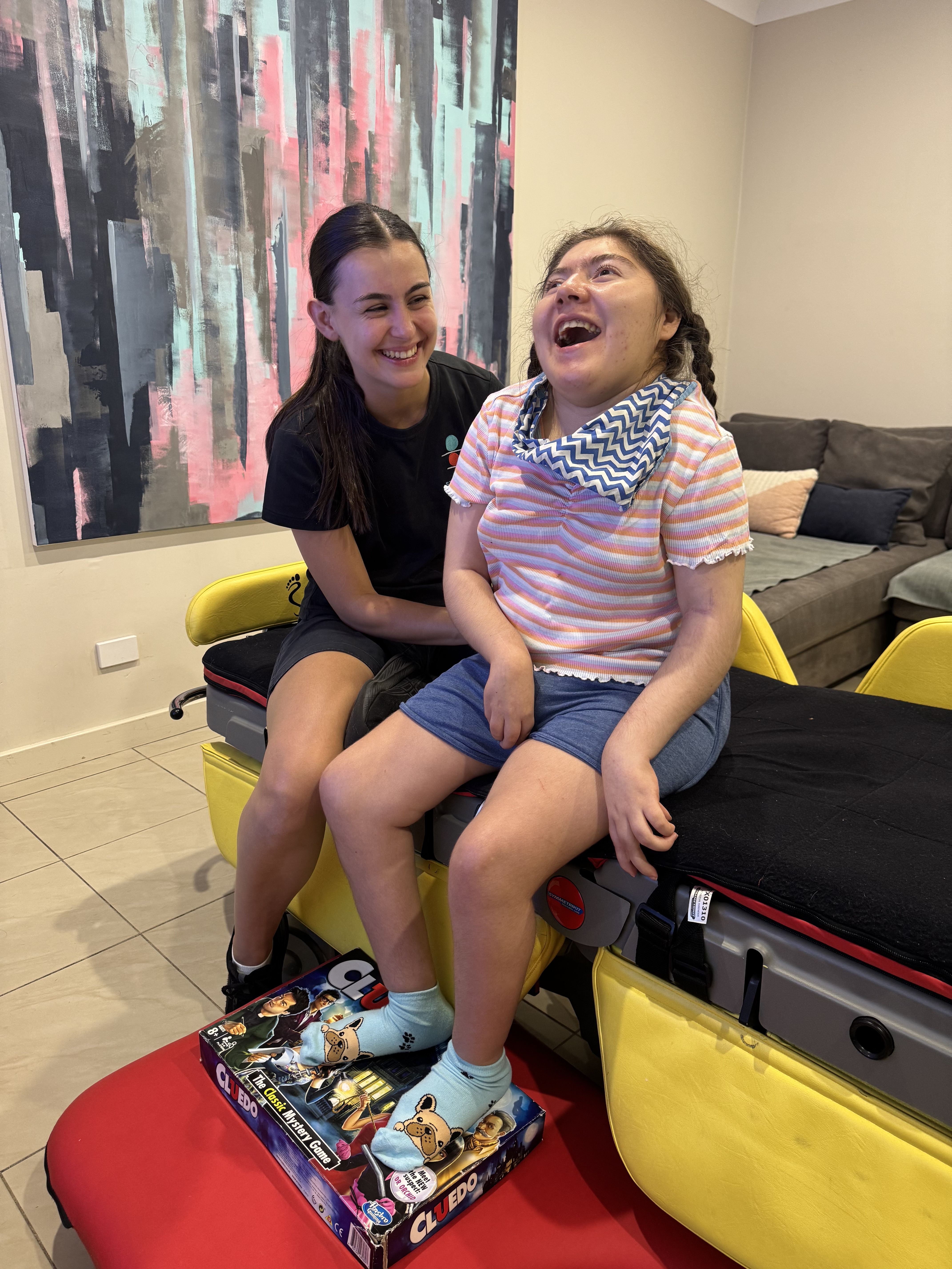 Amara sits upright on a moveable bed while a physiotherapist assists her. Amara's hair is pulled into two plaits and she's wearing a t-shirt, shorts, a neckerchief and socks with cute illustrated bulldogs printed on them. 
