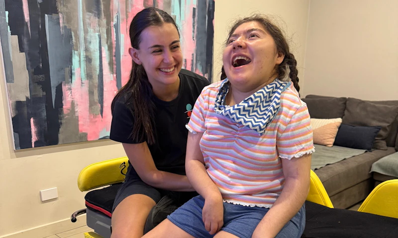 Amara sits upright on a moveable bed while a physiotherapist assists her. Amara's hair is pulled into two plaits and she's wearing a t-shirt, shorts, a neckerchief and socks with cute illustrated bulldogs printed on them.
