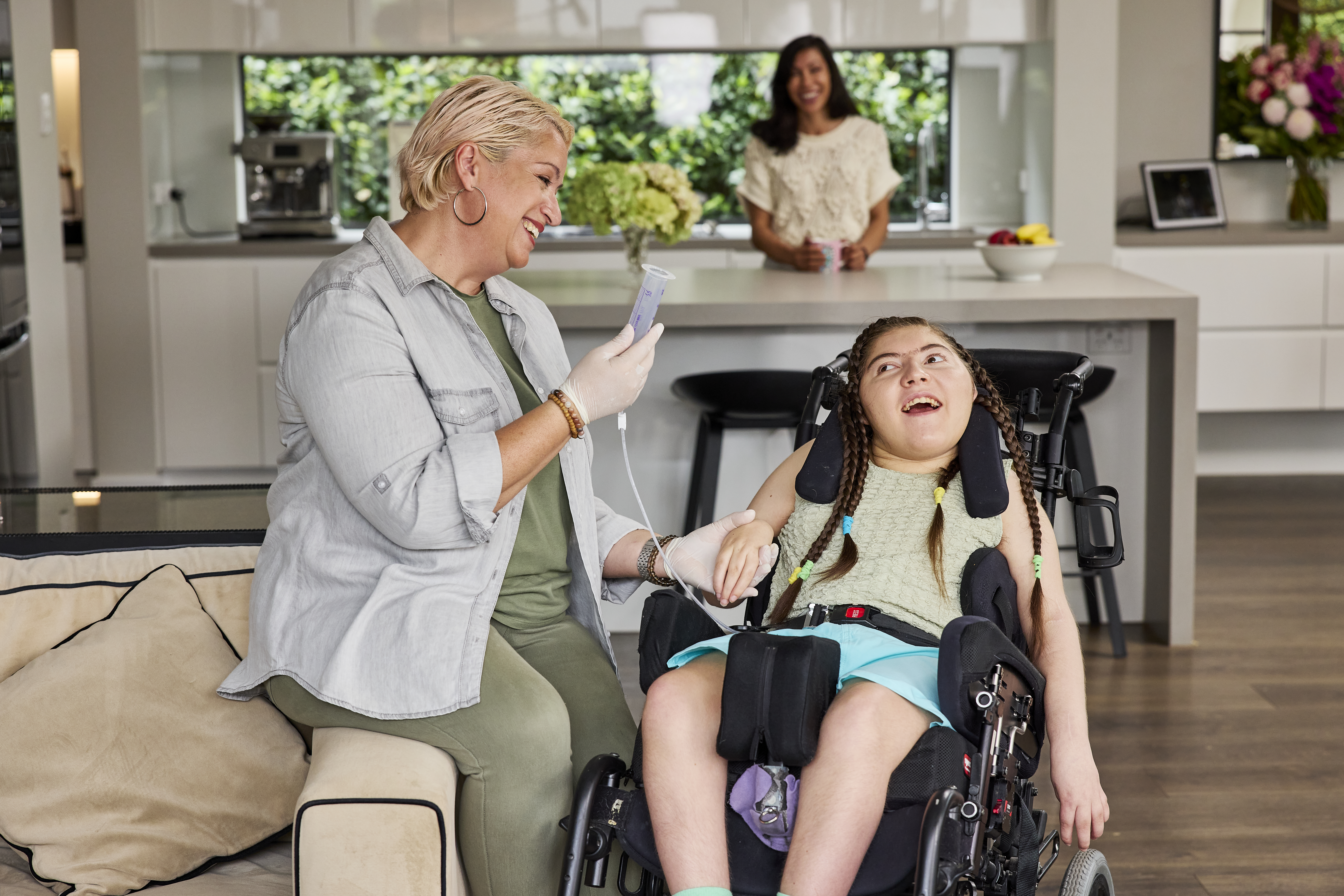 Amara, who is a wheelchair user, has long dark hair that's been plaited. She's in a lounge room with her support worker. Her Hireup support worker, who has short blonde hair and is dressed in khaki, perches on the arm of the couch, assisting Amara with her tube feeding. Amara's smiling at her support worker, while her mum watches on from the kitchen. 