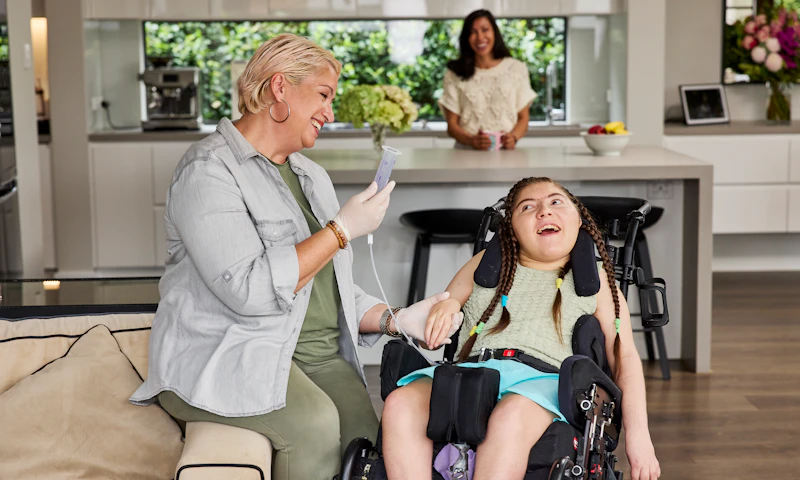 Amara, who is a wheelchair user, has long dark hair that's been plaited. She's in a lounge room with her support worker. Her Hireup support worker, who has short blonde hair and is dressed in khaki, perches on the arm of the couch, assisting Amara with her tube feeding. Amara's smiling at her support worker, while her mum watches on from the kitchen.