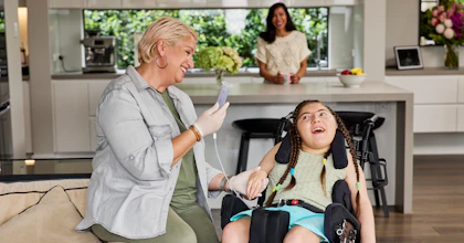 Amara, who is a wheelchair user, has long dark hair that's been plaited. She's in a lounge room with her support worker. Her Hireup support worker, who has short blonde hair and is dressed in khaki, perches on the arm of the couch, assisting Amara with her tube feeding. Amara's smiling at her support worker, while her mum watches on from the kitchen.