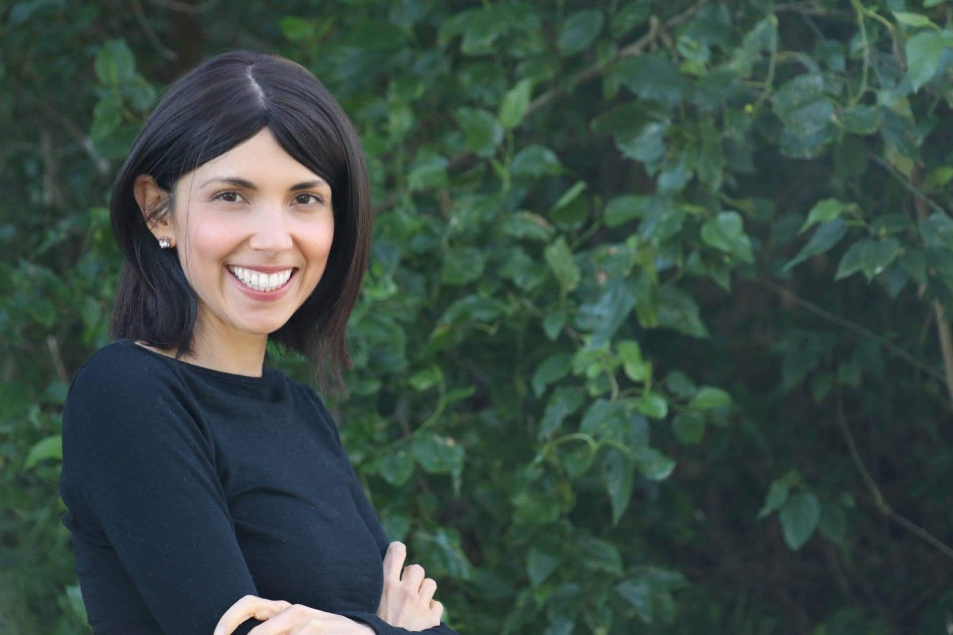 Natalia is a young woman with short, dark hair. She has her arms folded and is smiling. Natalia is standing in front of a vine. 