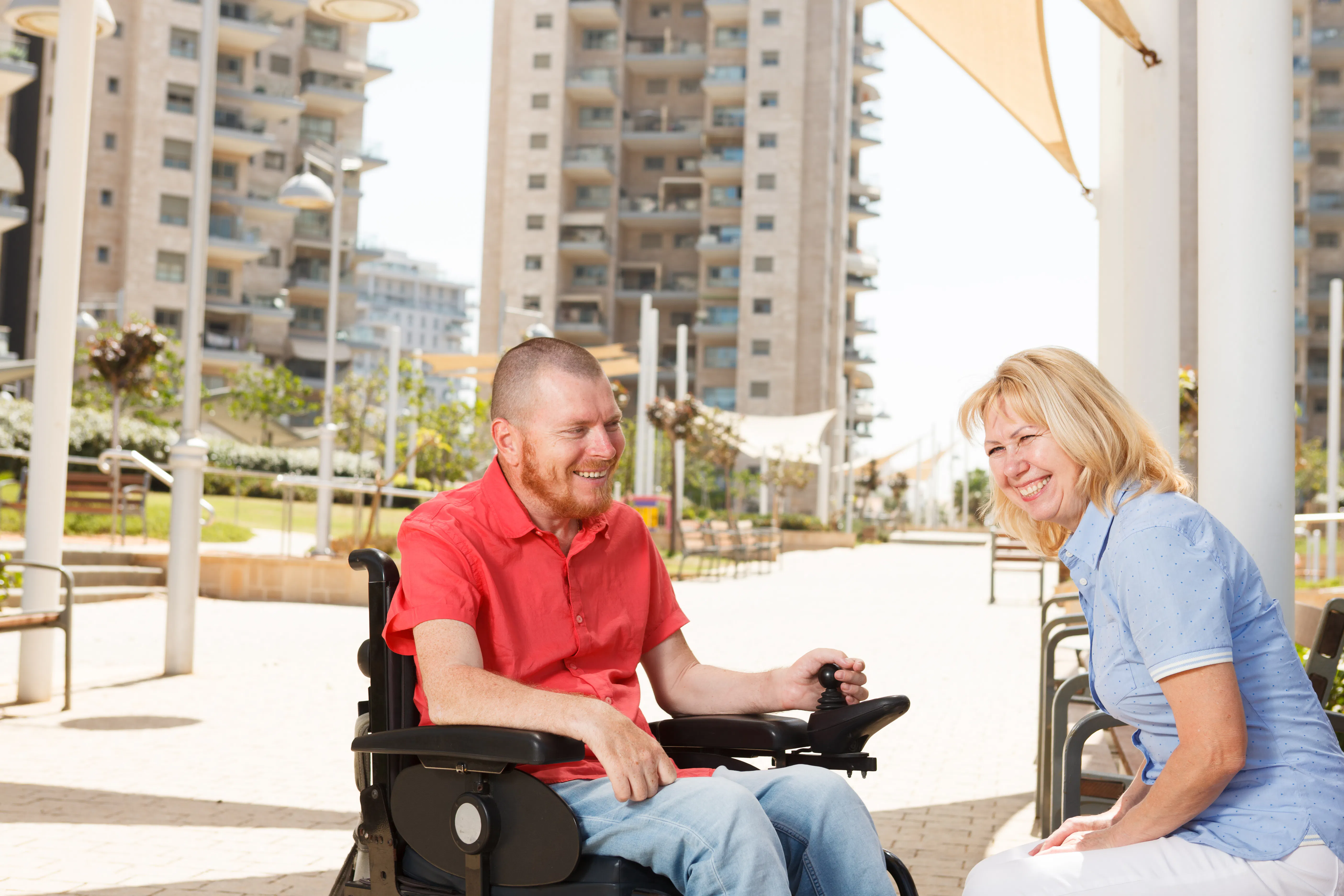 A man and a woman are sitting together smiling. The man has dark hair shaved short, and is wearing a red short sleeved collared shirt and light blue denim jeans. The man is a wheelchair user. The woman has shoulder length blonde hair and is wearing a pale blue collared shirt and white pants. There are apartment buildings and a park in the background.