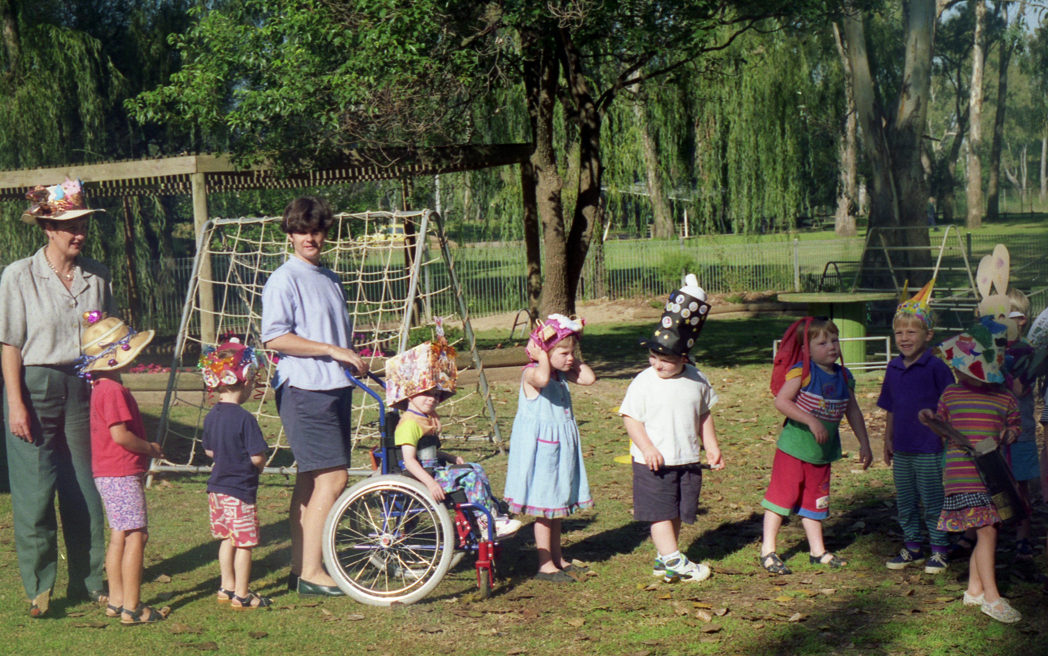 A line of school children wear festive hats in a sunny, green park accompanied by two adults. A young Carl Thompson features in the middle of the line in his wheelchair, wearing a large cardboard box creation on his head.