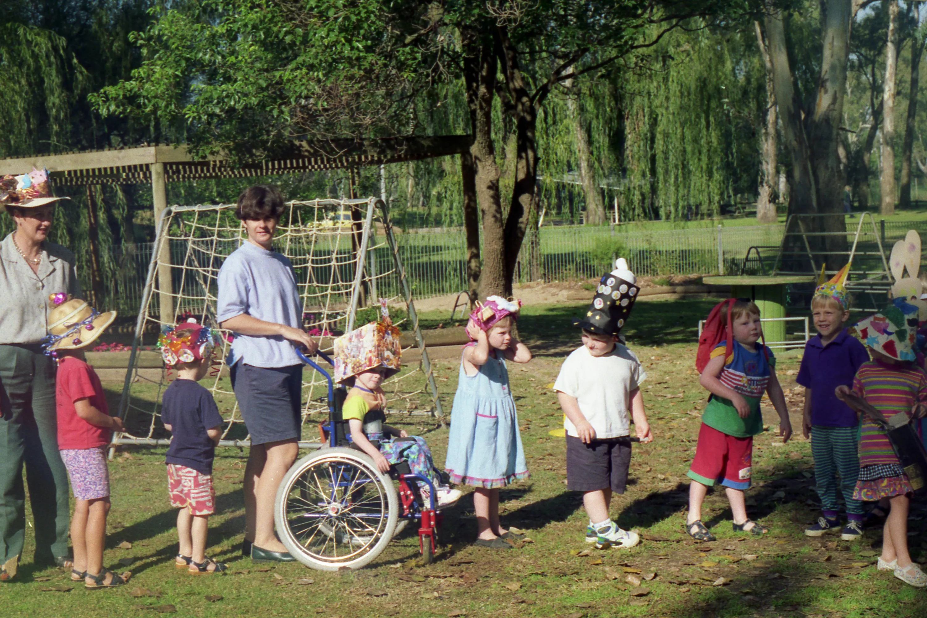 A line of school children wear festive hats in a sunny, green park accompanied by two adults. A young Carl Thompson features in the middle of the line in his wheelchair, wearing a large cardboard box creation on his head.