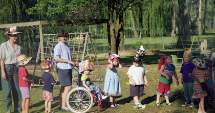 A line of school children wear festive hats in a sunny, green park accompanied by two adults. A young Carl Thompson features in the middle of the line in his wheelchair, wearing a large cardboard box creation on his head.