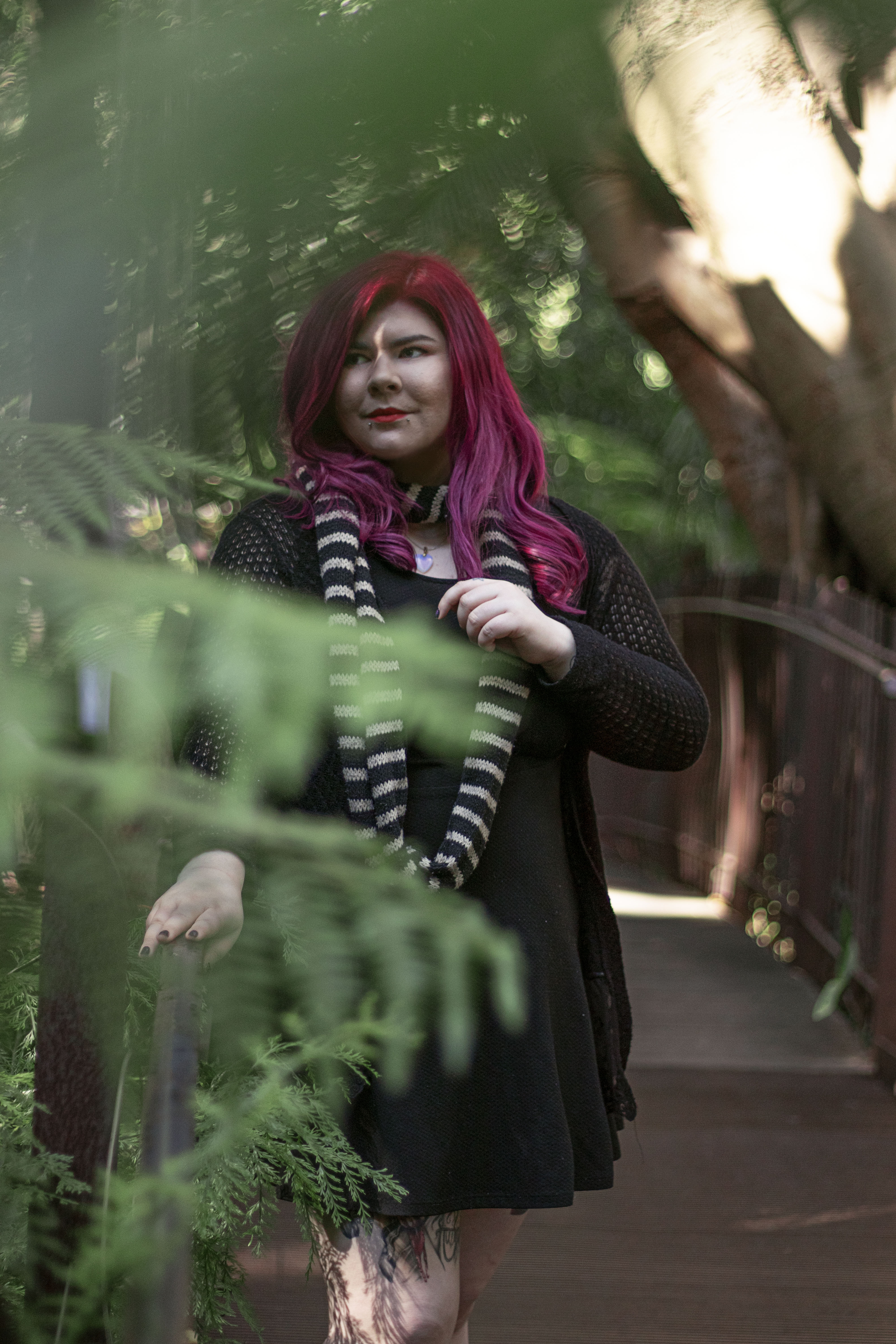 Zoe has long, pink hair and wears a black dress. She is standing on a bridge and is surrounded by trees. 