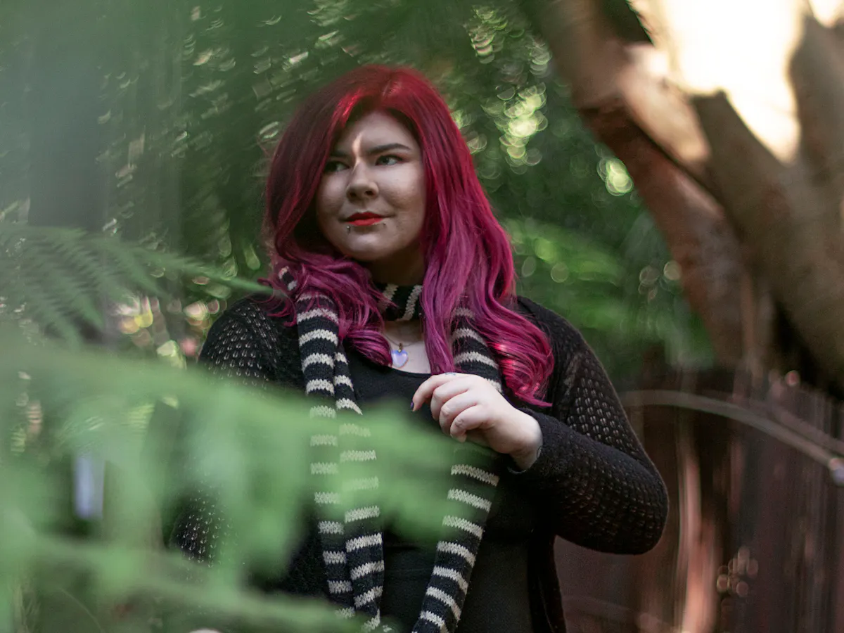 Zoe has long, pink hair and wears a black dress. She is standing on a bridge and is surrounded by trees.