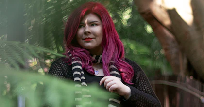 Zoe has long, pink hair and wears a black dress. She is standing on a bridge and is surrounded by trees.