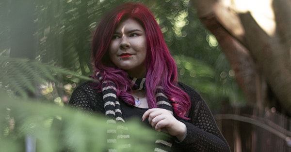 Zoe has long, pink hair and wears a black dress. She is standing on a bridge and is surrounded by trees.