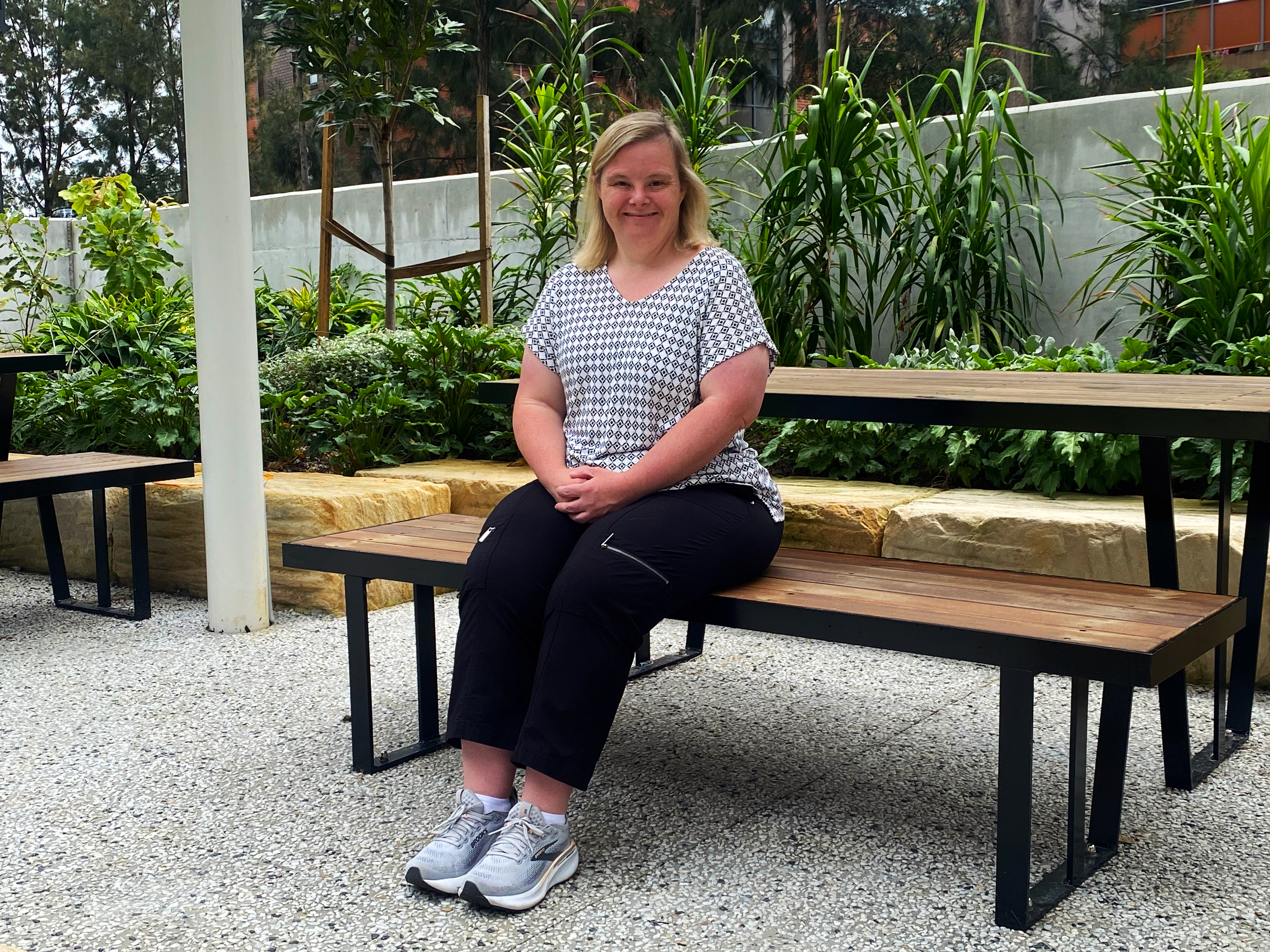 Tara, a Hireup client, sits on a picnic bench. She's dressed in a gingham-patterned top, black pants and joggers. Tara smiles gently with her hands clasped together in her lap.