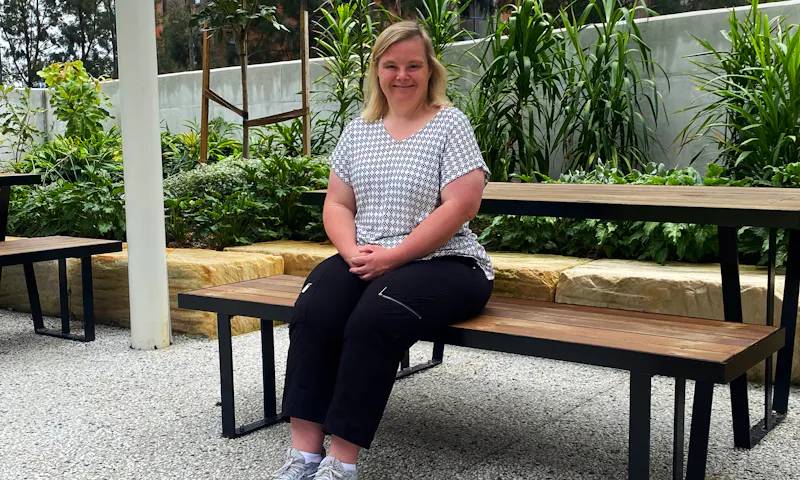Tara, a Hireup client, sits on a picnic bench. She's dressed in a gingham-patterned top, black pants and joggers. Tara smiles gently with her hands clasped together in her lap.