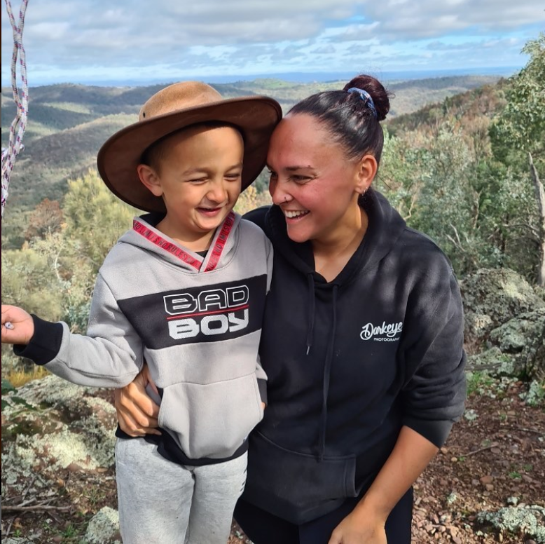 Brinae is kneeling and has her arm wrapped around her son. They're both laughing. Behind them is a valley of trees. 