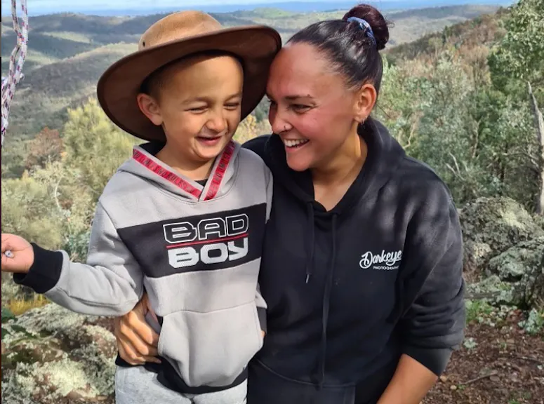 Brinae is kneeling and has her arm wrapped around her son. They're both laughing. Behind them is a valley of trees.