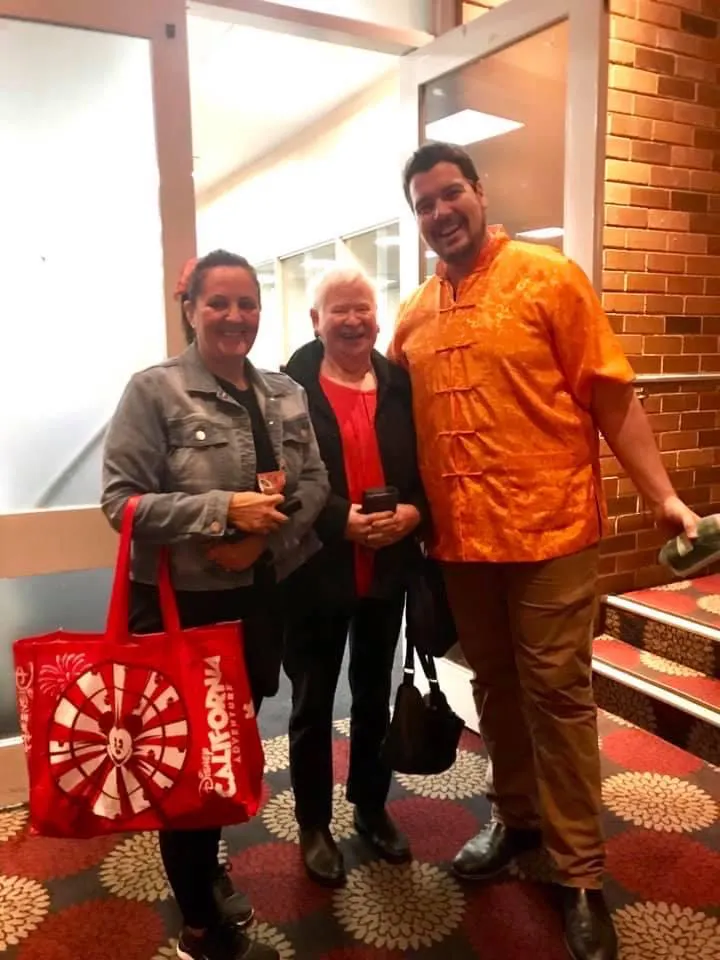 Noelene and Victoria are standing with Josh who is very tall and wearing a bright orange shirt. Victoria is hold a large red tote bag.