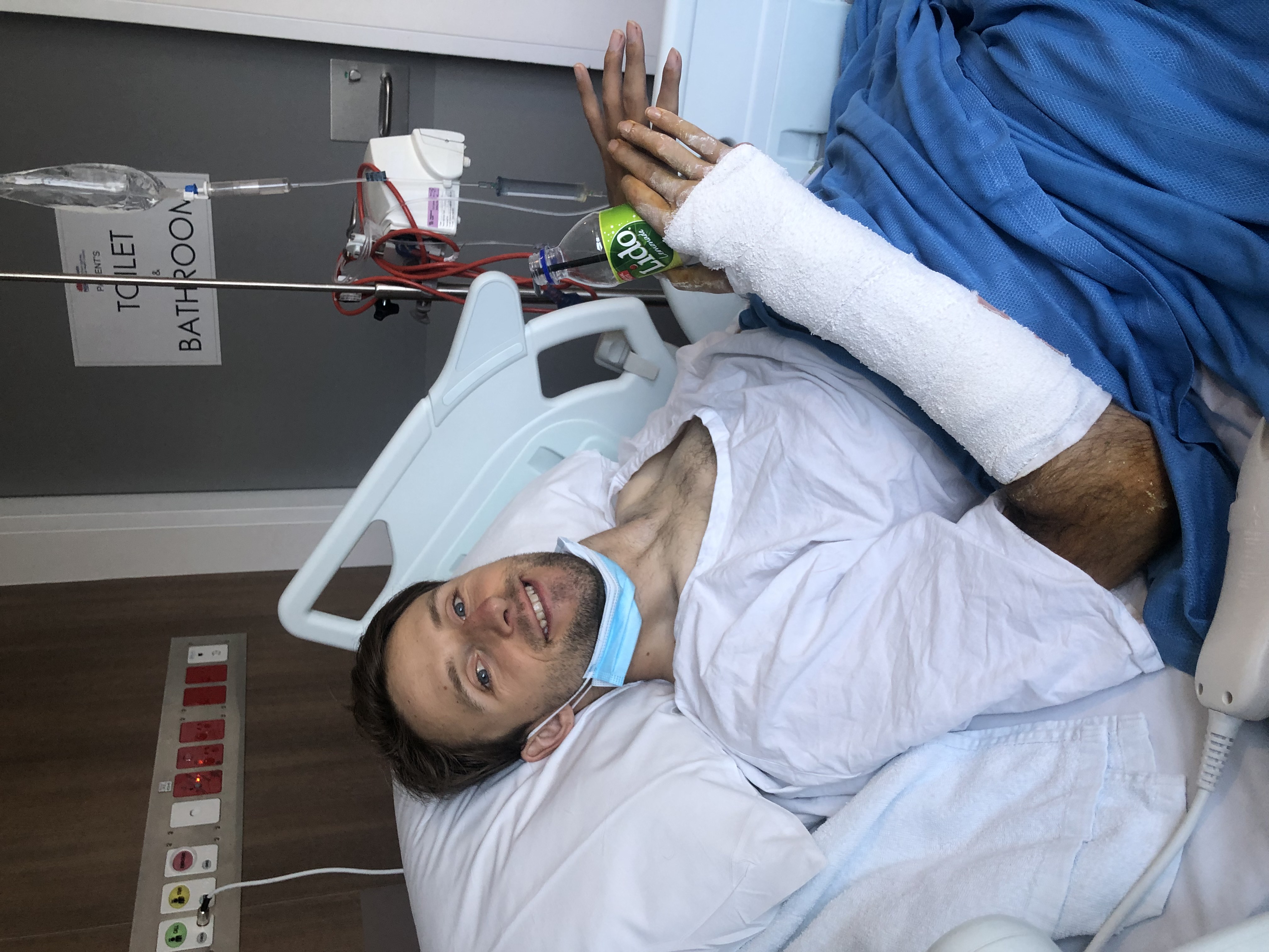 Steve Ralph is lying in a hospital bed. His right-arm is bandaged and he's holding a drink bottle. Steve is smiling. 
