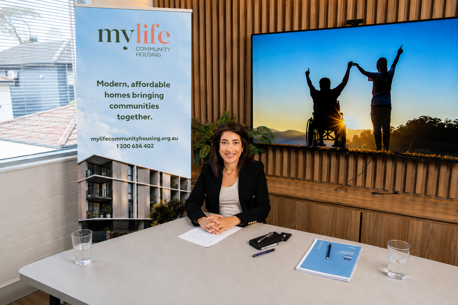 Karen Abdallah, the Managing Director and Founder of MyLife Community Housing, is seated at a desk. She has short dark hair, wears a dark coloured blazer, and smiles gently. A large 'MyLife Community Housing' banner is beside the desk. 