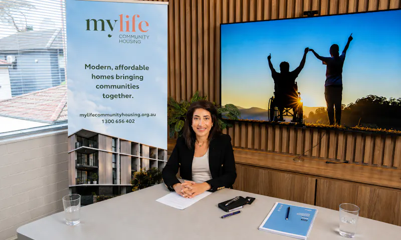 Karen Abdallah, the Managing Director and Founder of MyLife Community Housing, is seated at a desk. She has short dark hair, wears a dark coloured blazer, and smiles gently. A large 'MyLife Community Housing' banner is beside the desk.