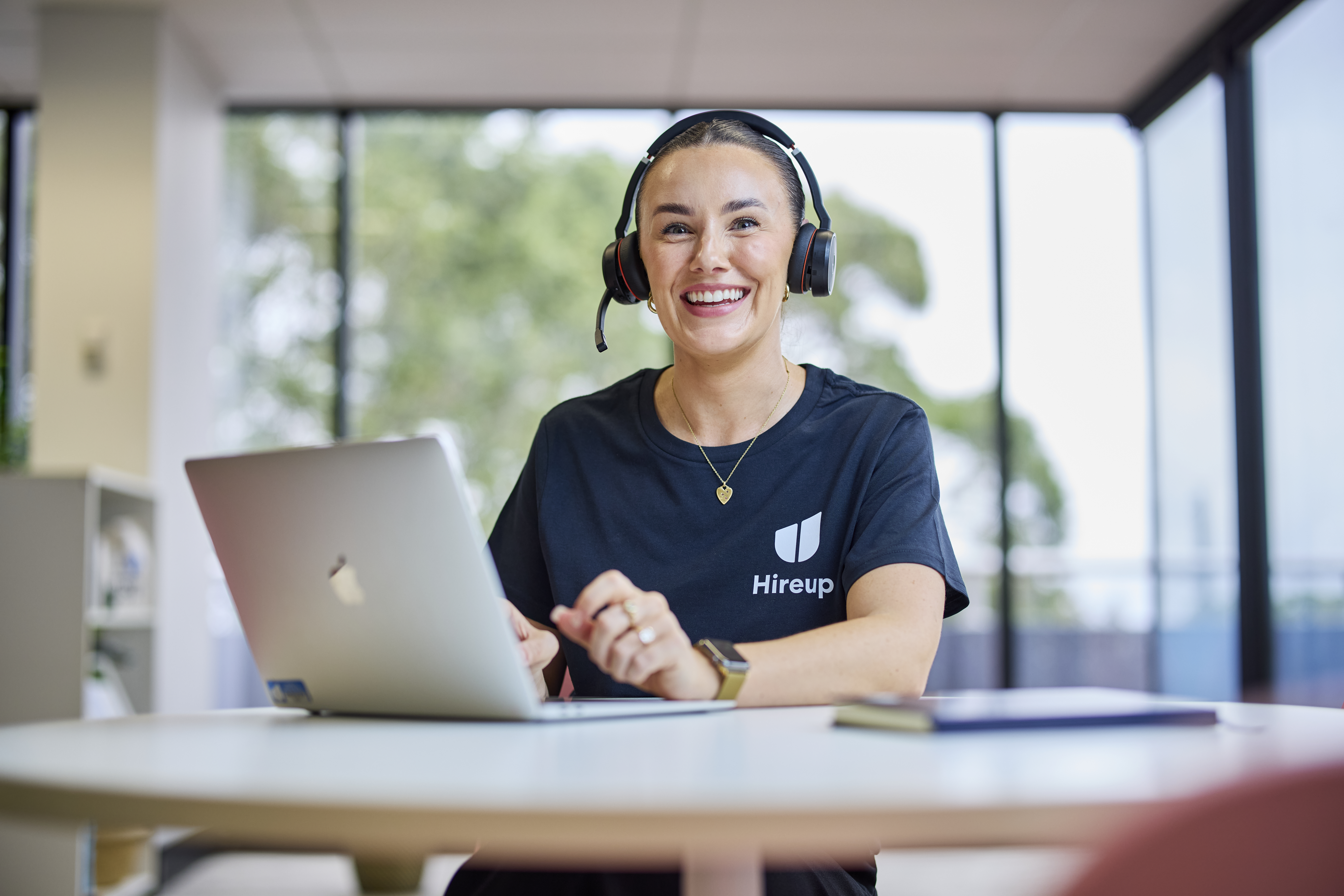 Shannon, a Hireup staff member, is sitting at a desk using a laptop. Shannon's wearing a Hireup t-shirt, wireless headphones and is smiling.