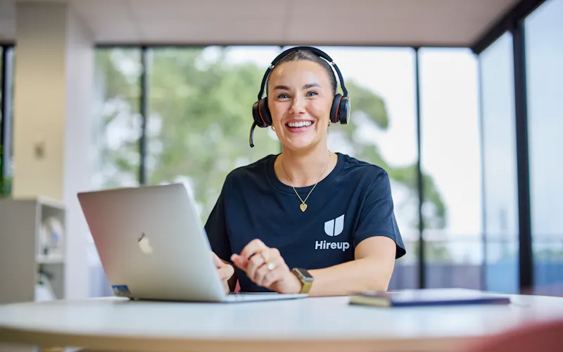 Shannon, a Hireup staff member, is sitting at a desk using a laptop. Shannon's wearing a Hireup t-shirt, wireless headphones and is smiling.