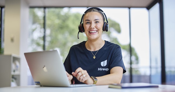 Shannon, a Hireup staff member, is sitting at a desk using a laptop. Shannon's wearing a Hireup t-shirt, wireless headphones and is smiling.