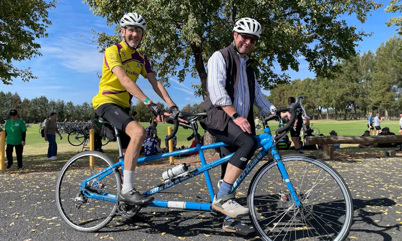 Andrew and his tandem partner, Leo, are at a park. They're wearing helmets, riding gear and are sitting on a blue tandem bike.