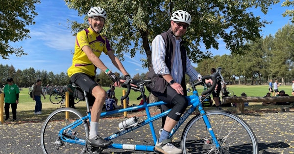 Andrew and his tandem partner, Leo, are at a park. They're wearing helmets, riding gear and are sitting on a blue tandem bike.