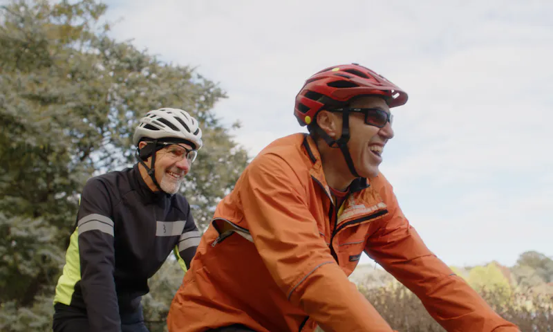 Andrew is riding a tandem bike with his Hireup support worker. They're both wearing helmets and jackets.
