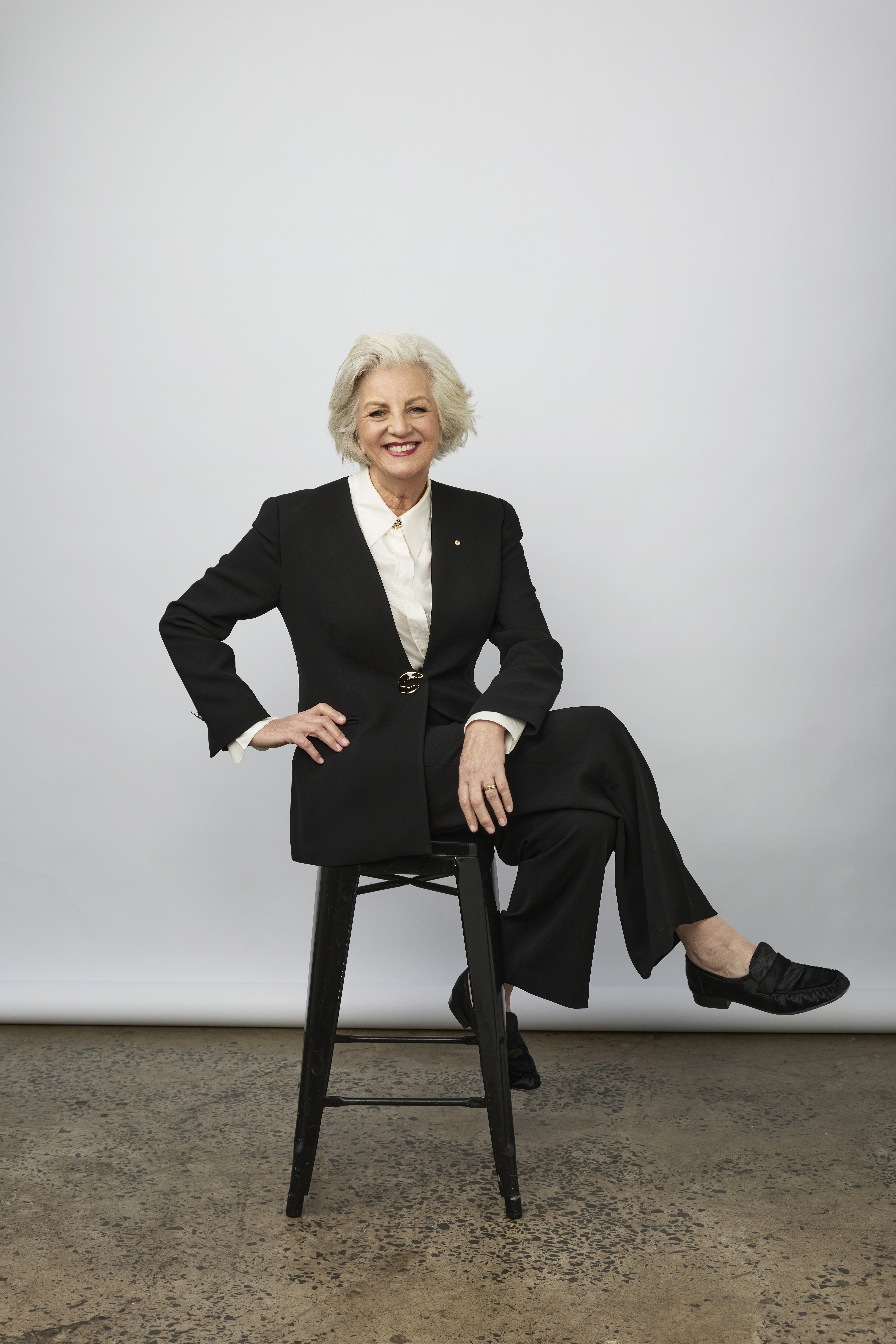 Jean Kittson sits cross-legged on a stool in front of a studio wall. She wears a black pantsuit and white shirt.