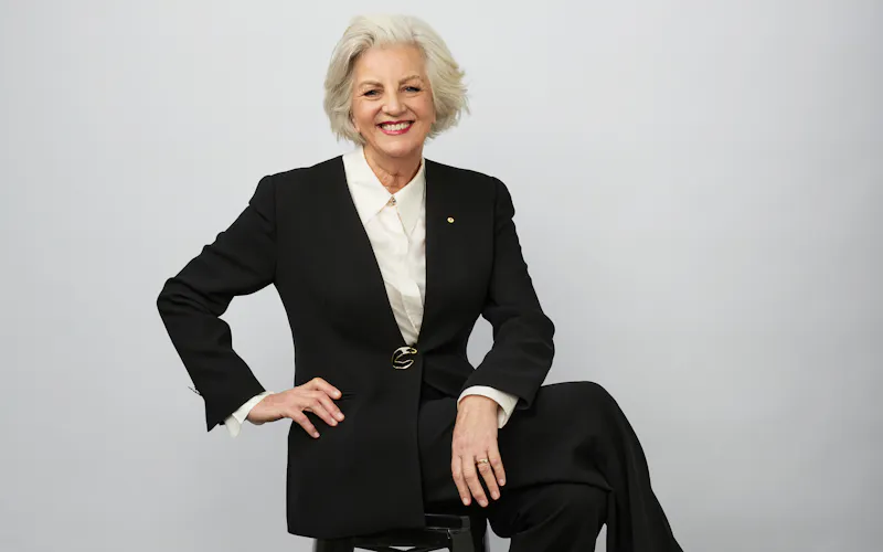 Jean Kittson sits cross-legged on a stool in front of a studio wall. She wears a black pantsuit and white shirt.