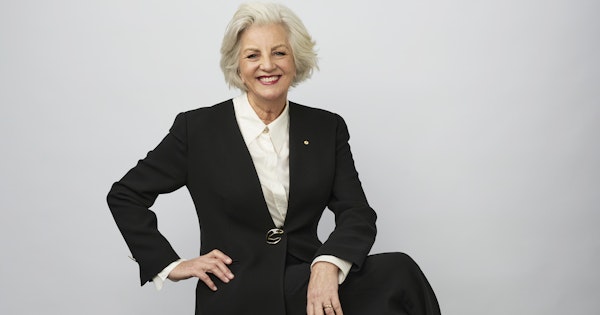 Jean Kittson sits cross-legged on a stool in front of a studio wall. She wears a black pantsuit and white shirt.