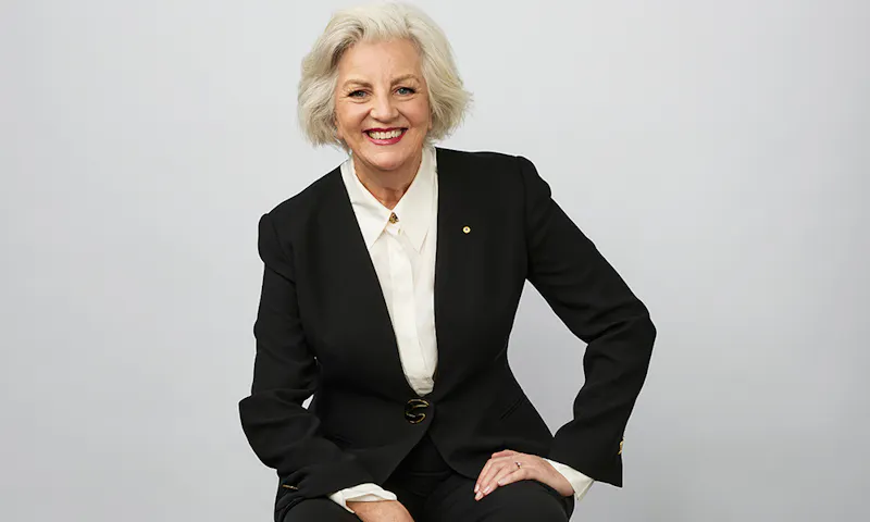 Jean Kittson is sitting on a stool. She has light grey hair and is wearing a black pant suit.