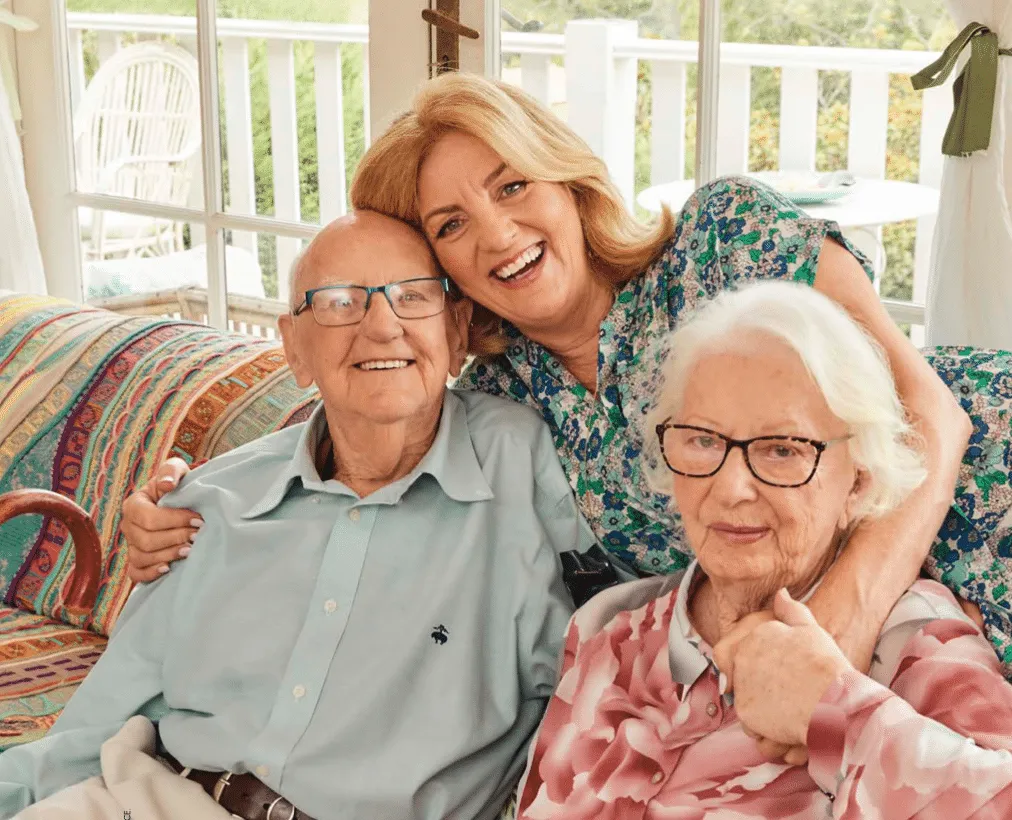 Jean Kittson is leaning over the back of a couch to hug her parents who are sitting on the couch. They're all smiling.