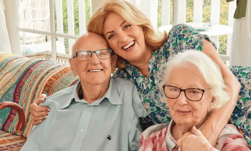 Jean Kittson is leaning over the back of a couch to hug her parents who are sitting on the couch. They're all smiling.