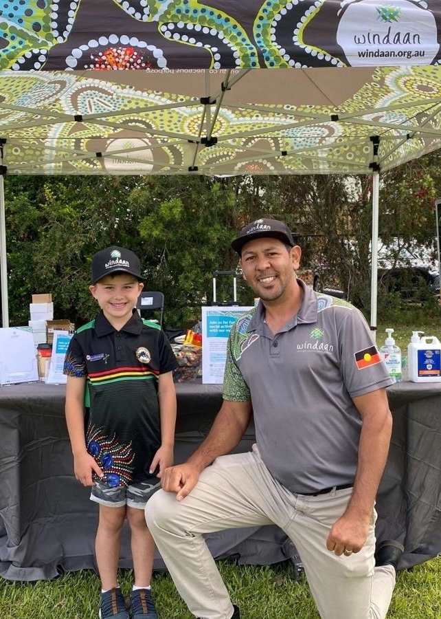 Joseph Archibald, manager of the Aboriginal Service Development at Windaan, kneels next to a young boy at an outdoor event. Both are smiling at the camera. 
