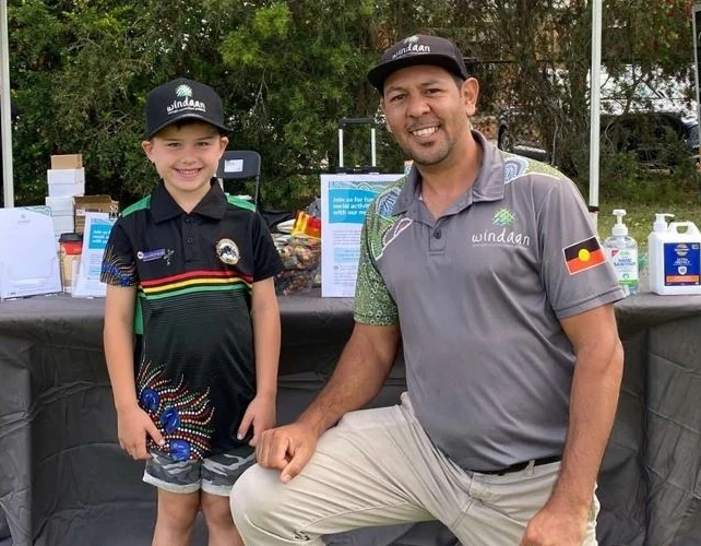 Joseph Archibald, manager of the Aboriginal Service Development at Windaan, kneels next to a young boy at an outdoor event. Both are smiling at the camera.