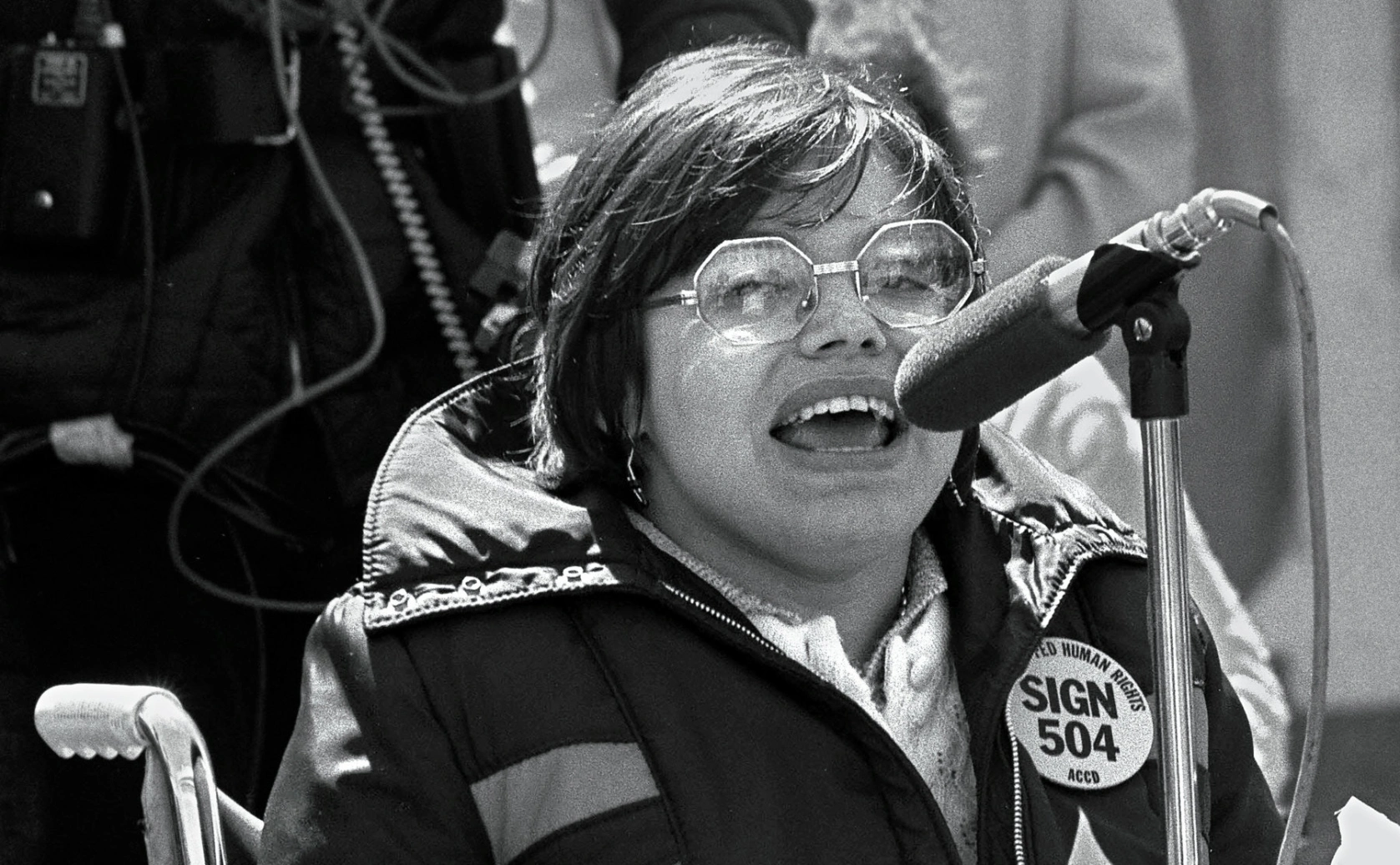 A black and white photo of Judy Heumann in 1977. Judy is a white woman with short brown hair who uses a wheelchair. She is wearing glasses and a jacket with a pin that says “Sign 504 Now” She is passionately speaking at a microphone.