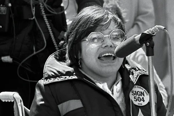A black and white photo of Judy Heumann in 1977. Judy is a white woman with short brown hair who uses a wheelchair. She is wearing glasses and a jacket with a pin that says “Sign 504 Now” She is passionately speaking at a microphone.