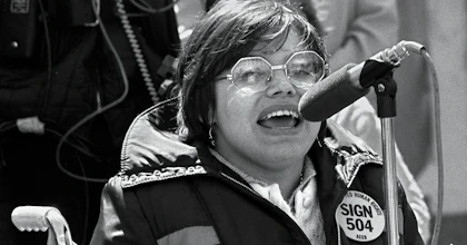 A black and white photo of Judy Heumann in 1977. Judy is a white woman with short brown hair who uses a wheelchair. She is wearing glasses and a jacket with a pin that says “Sign 504 Now” She is passionately speaking at a microphone.