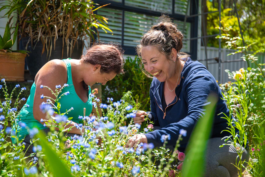 Portia and Sam are both knelt down in the garden. They are both cracking up laughing while picking flowers.