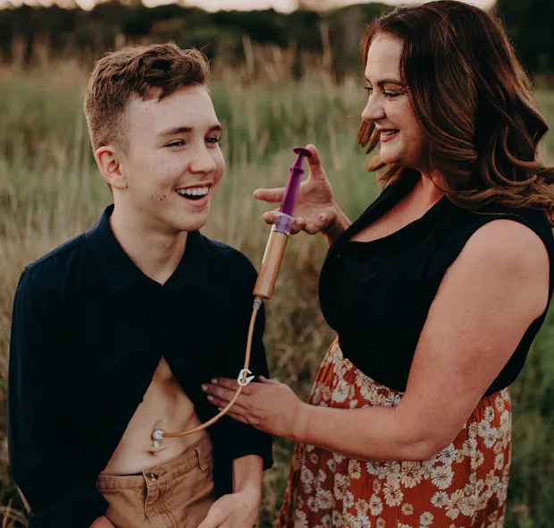 Sarah Thomas, a woman with dark, wavy hair, is supporting her son, Lewis, with his tube-feeding. They're smiling and are standing in a field of tall grass.