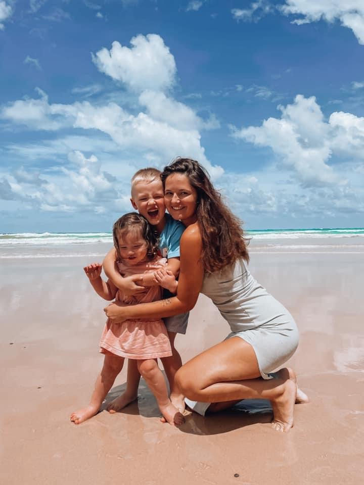 Angel is crouched down and is cuddling her two children. They're on a beach. The sea, sand and sky is bright with sunshine.