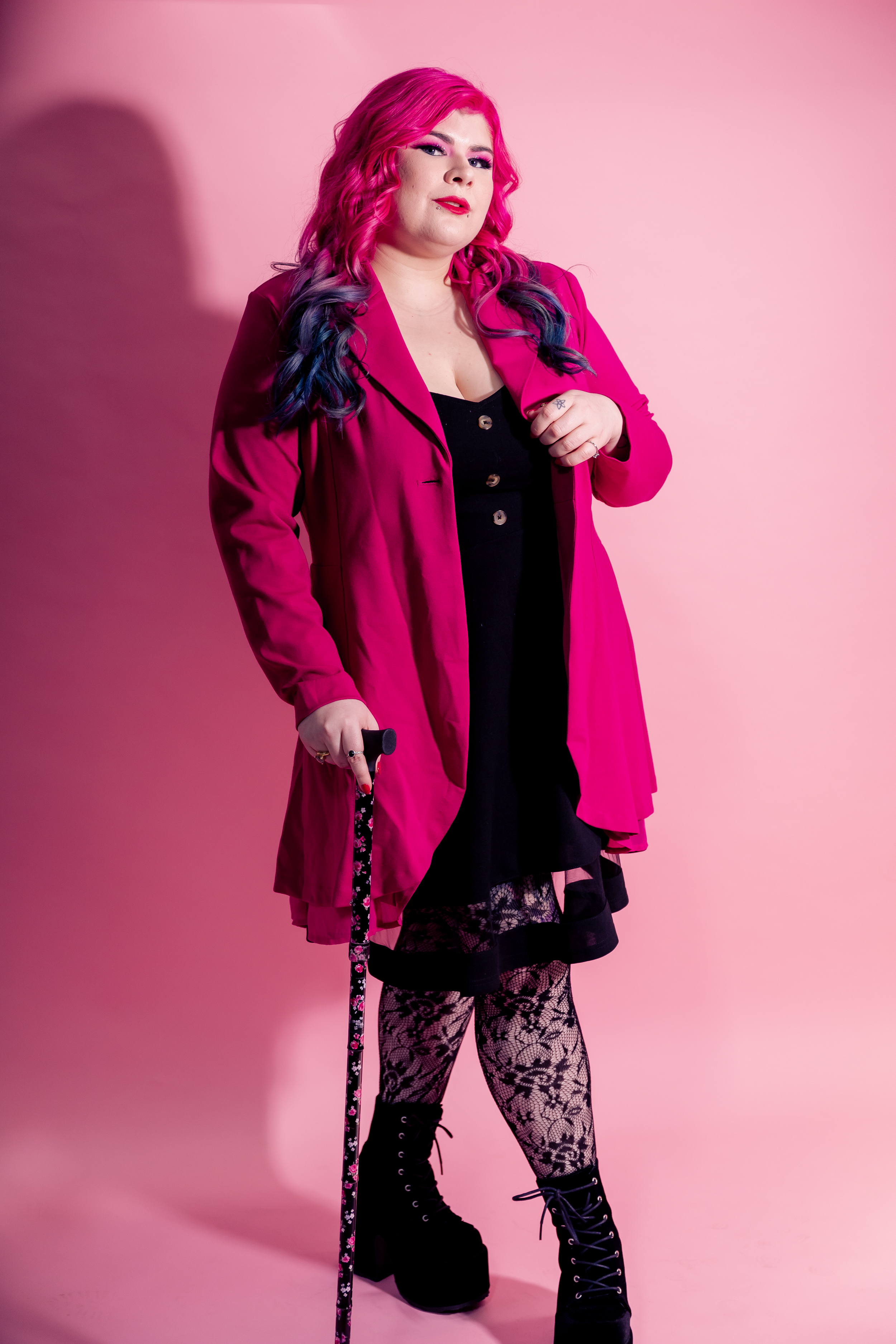 Zoe Simmons, a disability advocate and writer, is wearing a bright pink coat, a black dress, floral fishnet stockings, and black heeled ankle boots. She has pale skin, long pink and blue hair, and bright makeup. She’s standing against a pink backdrop and holding a cane. 