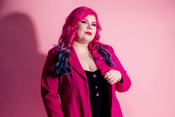 Zoe Simmons, a disability advocate and writer, is wearing a bright pink coat, a black dress, floral fishnet stockings, and black heeled ankle boots. She has pale skin, long pink and blue hair, and bright makeup. She’s standing against a pink backdrop and holding a cane.