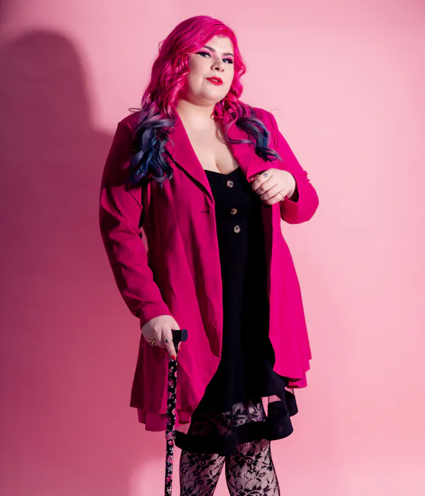 Zoe Simmons, a disability advocate and writer, is wearing a bright pink coat, a black dress, floral fishnet stockings, and black heeled ankle boots. She has pale skin, long pink and blue hair, and bright makeup. She’s standing against a pink backdrop and holding a cane.