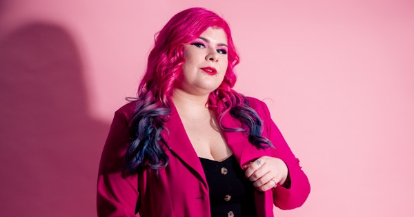 Zoe Simmons, a disability advocate and writer, is wearing a bright pink coat, a black dress, floral fishnet stockings, and black heeled ankle boots. She has pale skin, long pink and blue hair, and bright makeup. She’s standing against a pink backdrop and holding a cane.