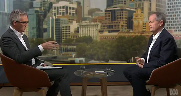 Two men in suits sit a across a glass table. The one on the left, in glasses, is talking.